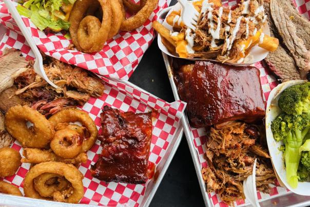 An overhead view of two trays piled with a variety of barbecue, including ribs, pulled pork, brisket, onion rings, and broccoli, on red-and-white checkered paper liners in Morehead, KY.