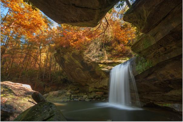 A scenic waterfall cascading into a calm pool of water, viewed from beneath a large, mossy rock overhang. The surrounding forest is filled with vibrant autumn foliage in brilliant shades of orange, yellow, and red, creating a colorful canopy above the gorge in Morehead, KY.
