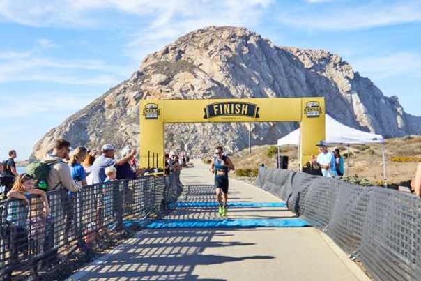 Man running across the finish line at the IRONMAN 70.3 lined with clapping spectators, with Morro Rock in the background.