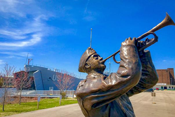 Bronze statue of ww2 soldier playing trumpet with ww2 landing ship docked in distance