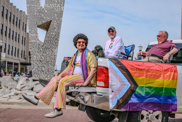 Three individuals on a decorated float featuring a rainbow pride flag, participating in a public event. One person sports dark curly hair and colorful attire, while sitting beside others in casual wear. Modern sculpture visible in the background.