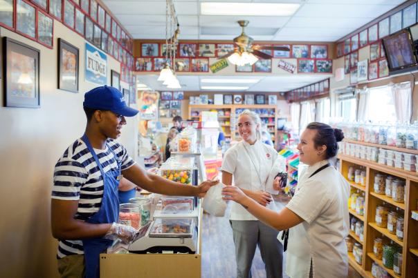 employee in blue and white striped shirt, blue apron and hat waiting on two people in candy store