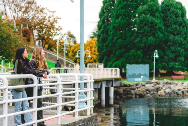 Two people pause along Nanaimo’s waterfront walkway, leaning on a railing and enjoying the view of the calm water and nearby park.