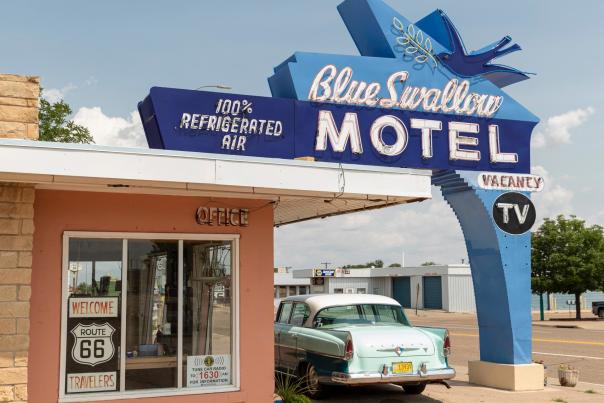 A classic 50's car sits beneath an unlit neon sign that reads "Blue Swallow Motel, 100% Refrigerated Air, Vacancy, TV"