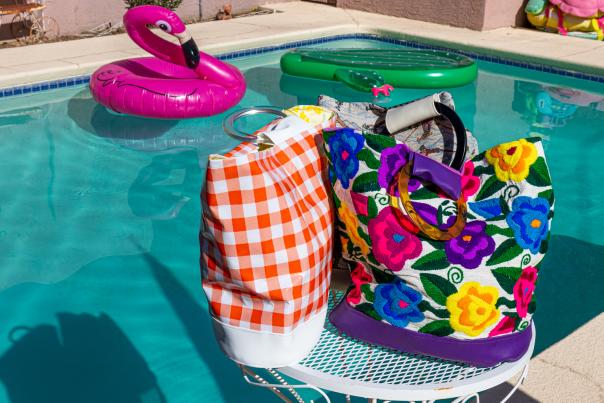 A colorful poolside scene featuring a vibrant, floral-patterned bag and a checked towel on a white metal table, with inflatable pool rings floating in the background.