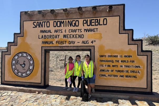 A group of three girls poses for a picture with the Santo Domingo Pueblo welcome sign after completing a litterl cleanup event.