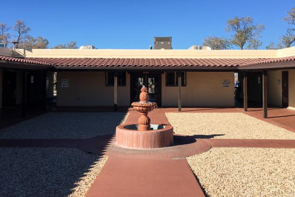 A view of a courtyard featuring a central tiered fountain, surrounded by a wide pebbled area and a building with a terracotta-colored tiled roof under a clear blue sky.