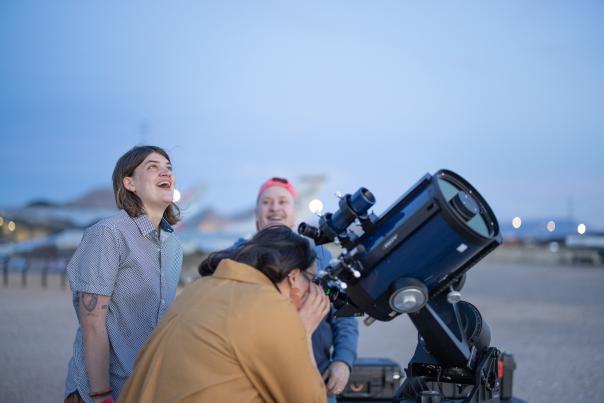 a person is hunched over looking through a large black telescope as two people stand in the background smiling