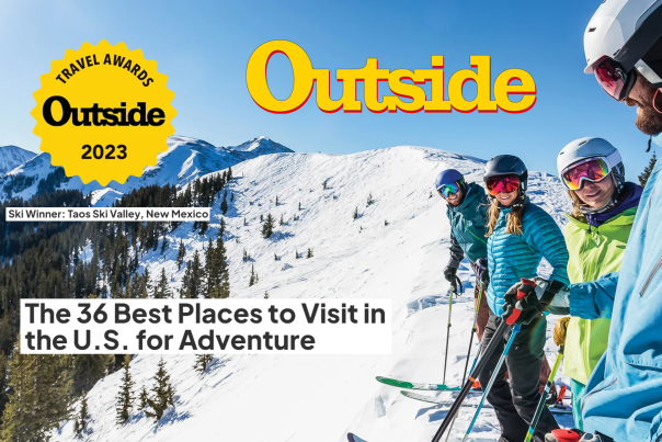Four skiers clad in winter gear stand on the peak of a snowy basin, accented by green trees. Text reads Outside, Travel Awards 2023. Ski Winner: Taos Ski Valley, New Mexico. The 36 Best Places to Visit In the U.S. for Adventure