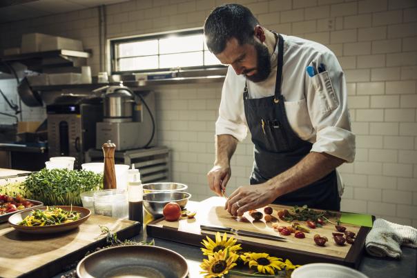 A chef diligently prepares food in a professional kitchen. The countertop is adorned with various fresh ingredients, including herbs and vegetables.