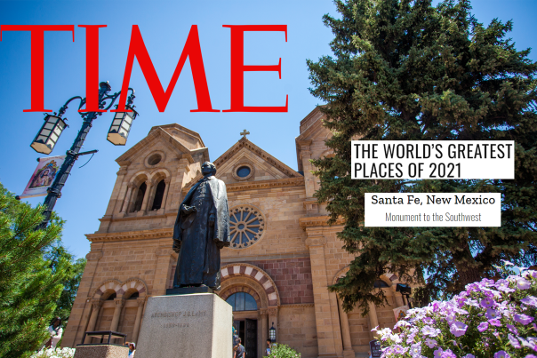 A statue of a priest stands before a tan chapel, surrounded by green trees and pink bushes of flowers. Text reads TIME, The Worlds Greatest Places of 2021. Santa Fe New Mexico. Monument to the Southwest.