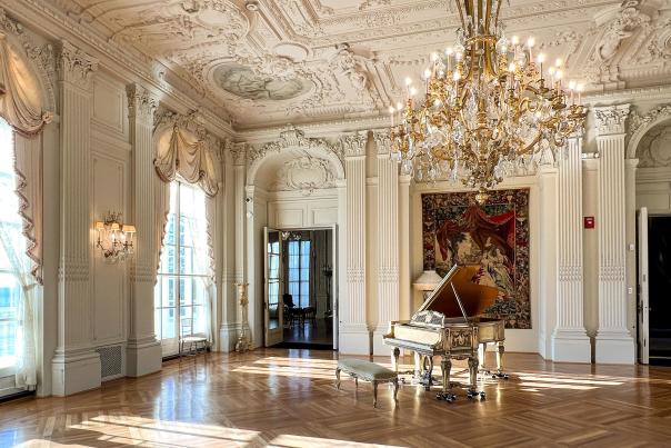 Detailed interior of Rosecliff Mansion's ballroom, including gold and crystal chandelier hanging in the middle of the room. There is an exquisite place piano on the far side of the room, and behind there is a historic art tapestry. Light comes through the two windows on the left, and fills the inside with sparkling light. Carved figures and designs cover the walls, columns, and ceilings.
