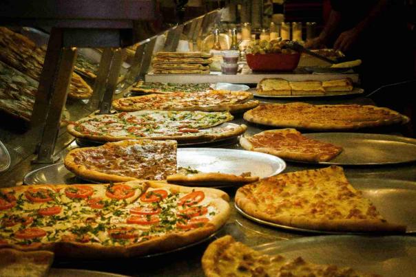 Fresh New York pizza slices displayed behind the counter at a busy Manhattan pizzeria with classic thin-crust and Sicilian-style pies.