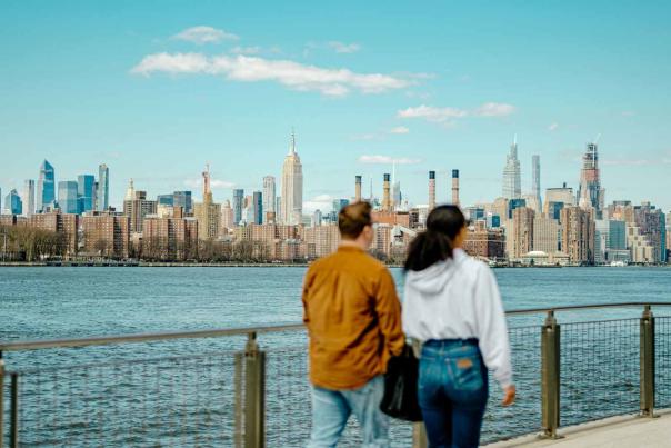 Two people standing along a waterfront promenade overlooking the Manhattan skyline on a bright day with the Empire State Building visible across the river.