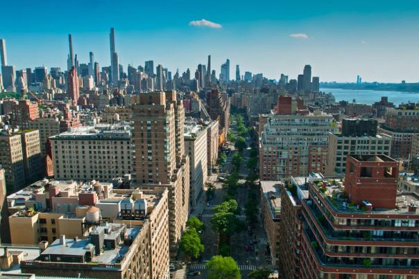 Aerial drone photograph looking north along Broadway through upper Manhattan, with tree-lined medians flanked by mid-rise residential and commercial buildings in the foreground, and the dense Midtown Manhattan skyline rising against a clear blue sky in the background.