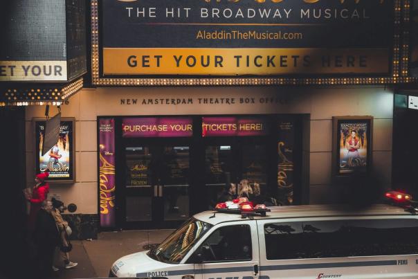 The New Amsterdam Theatre box office on 42nd Street in New York City displaying the Disney Aladdin Broadway musical marquee at night with an NYPD vehicle in the foreground