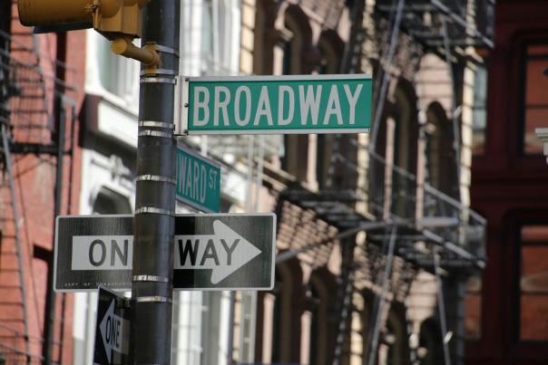 Green Broadway street sign on a pole with a one way sign and historic brick buildings with fire escapes in the background in New York City
