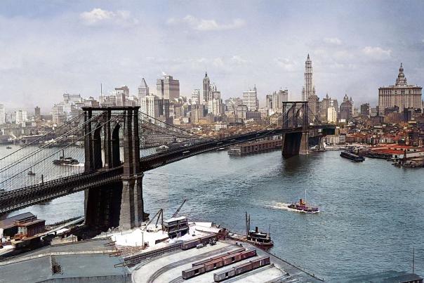 Colorized early 20th-century aerial photograph of the Brooklyn Bridge spanning the East River, with the Lower Manhattan skyline — including the Woolworth Building and Municipal Building — rising behind it, and tugboats and waterfront rail yards visible in the foreground.