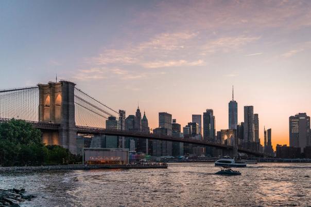Brooklyn Bridge with the Manhattan skyline glowing behind it at sunset, viewed from the water in New York City