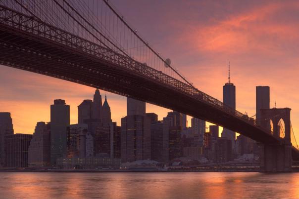 Brooklyn Bridge silhouetted against a vivid red and orange sunrise sky, with the Manhattan skyline reflected across the calm East River below