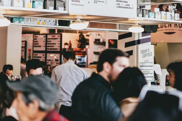 A crowded coffee shop counter with a barista working behind the bar and customers gathered in front, menu boards and bagged coffee beans visible on the shelves above