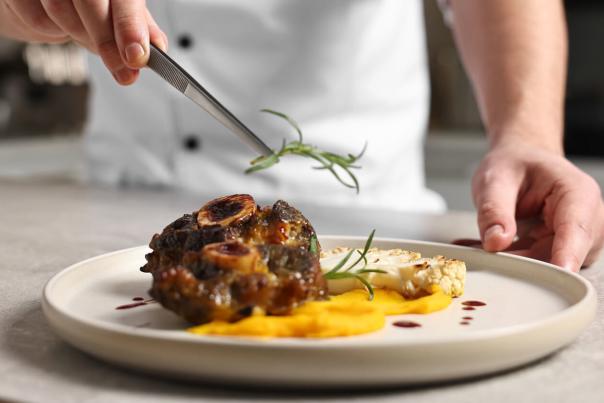 Chef using precision tweezers to place a rosemary sprig on a braised meat course over golden purée on a ceramic plate