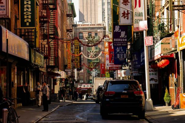 People and cars moving through a busy Chinatown street in New York City lined with storefronts and signs