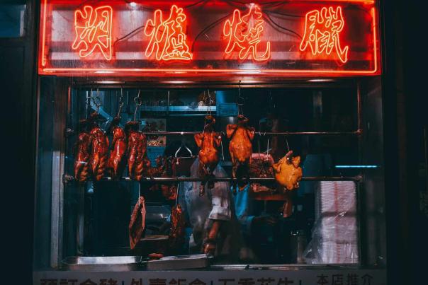 Roasted ducks and poultry hanging in a Chinatown shop window beneath a glowing red neon sign with Chinese characters