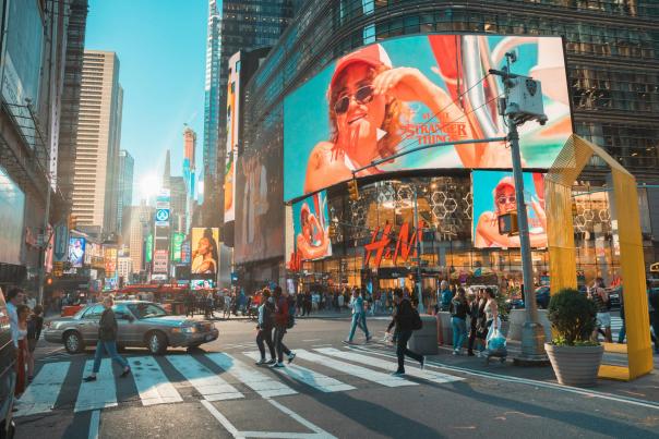 Busy daytime intersection in Times Square, New York City, with pedestrians crossing, large digital billboards, and the H&M storefront surrounded by tall buildings.