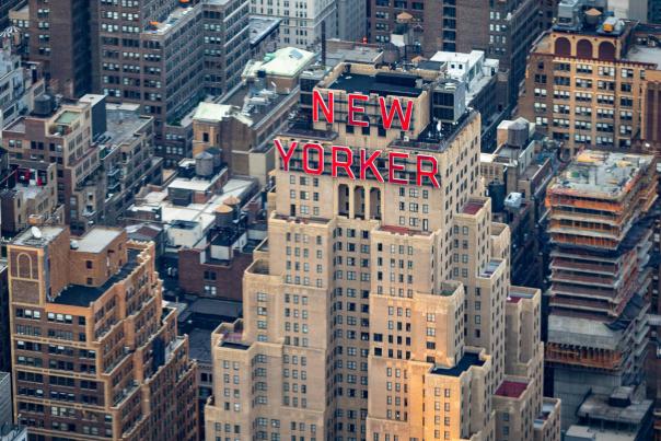 Aerial view of the New Yorker Hotel with its iconic red rooftop sign surrounded by dense Midtown Manhattan skyscrapers in New York City.