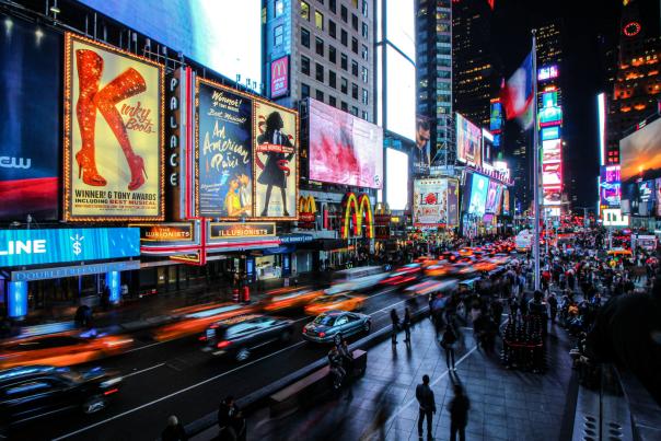 Nighttime view of Times Square in New York City with bright theater billboards, heavy traffic with light streaks, and crowds walking along the sidewalks.