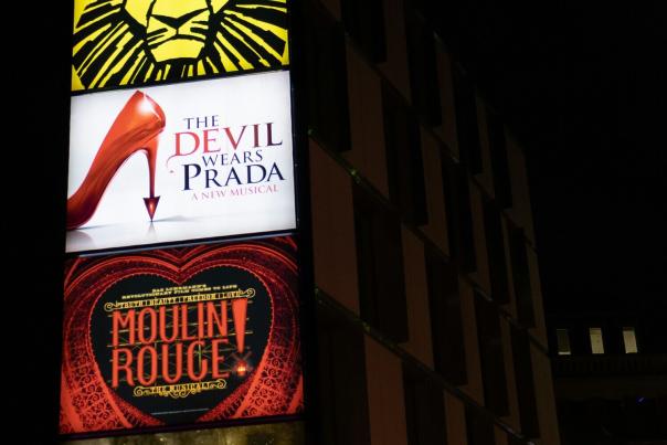 Illuminated theatre billboard tower at night showing Hadestown at the bottom alongside Hercules, The Lion King, The Devil Wears Prada, and Moulin Rouge