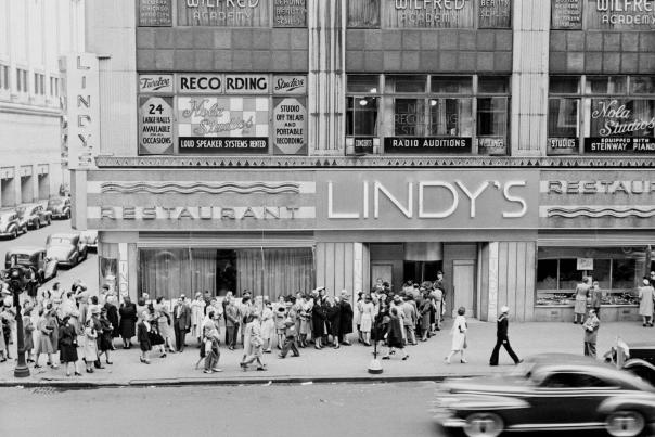 Crowds lined up outside Lindy's Restaurant on a busy Midtown Manhattan street in the 1940s with vintage cars passing below