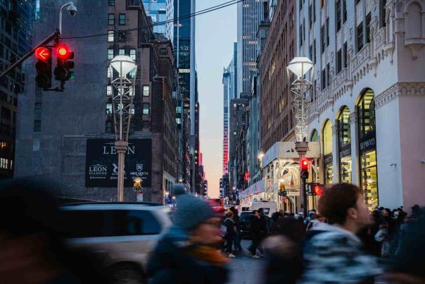 Pedestrians crossing a busy Midtown Manhattan intersection at dusk with skyscrapers and red traffic lights overhead