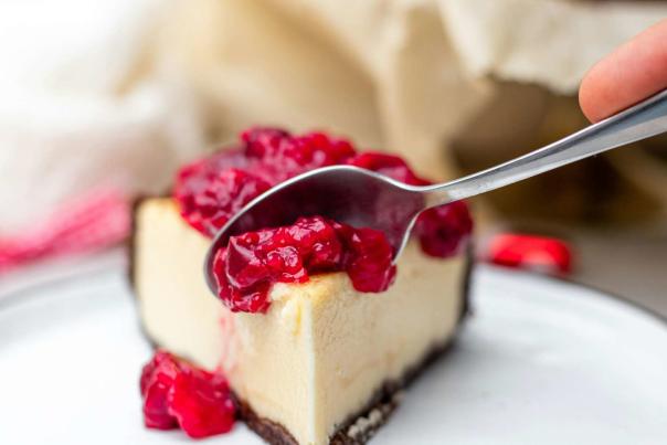 Slice of New York cheesecake with chocolate cookie crust and sour cherry topping on a plate, capturing the dense creamy texture that defines the style