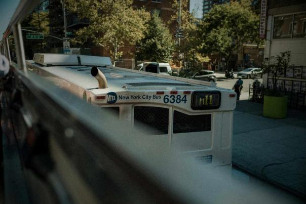 MTA New York City bus traveling through Manhattan streets during daylight with surrounding neighborhood buildings and traffic visible.