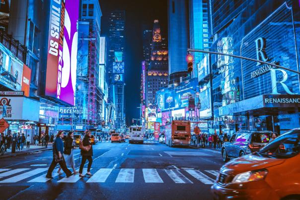 Busy nighttime scene in Times Square, New York City, with colorful digital billboards, traffic, pedestrians crossing the street, and bright city lights illuminating the avenue.