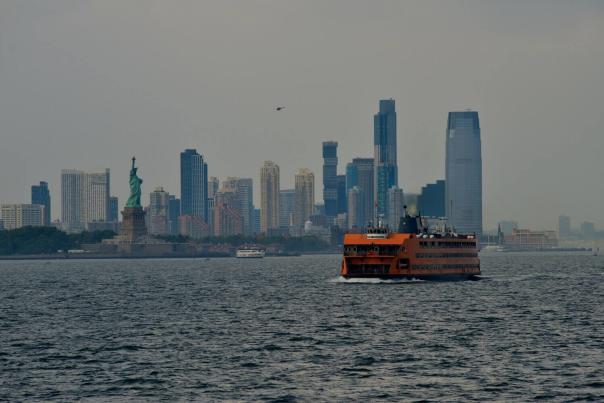 The Staten Island Ferry crosses New York Harbor on an overcast day, with the Statue of Liberty and Jersey City skyline visible in the background