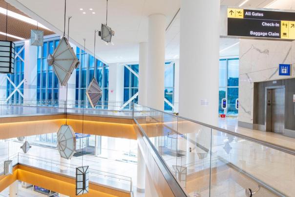 Interior of a modern Newark Liberty International Airport terminal featuring a bright, multi-level atrium with geometric diamond-shaped hanging sculptures, glass railings, white marble walls, and overhead directional signage pointing to All Gates, Check-In, and Baggage Claim.