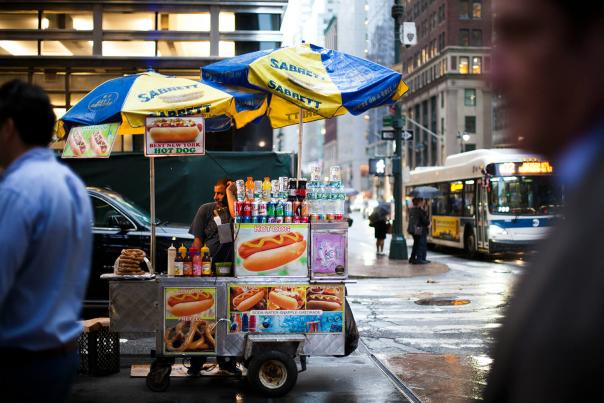 A hot dog vendor cart stationed at a busy New York City intersection surrounded by pedestrians
