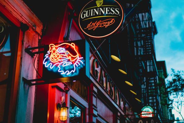 Exterior of an Irish pub in New York City at dusk, featuring a glowing Guinness sign above a neon crab logo and a red-painted facade with fire escapes in the background.