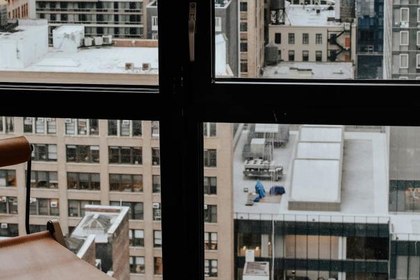 Floor-to-ceiling hotel room window with dark frames overlooking a dense Midtown Manhattan skyline on a grey winter day, with the corner of a leather chair visible in the foreground.