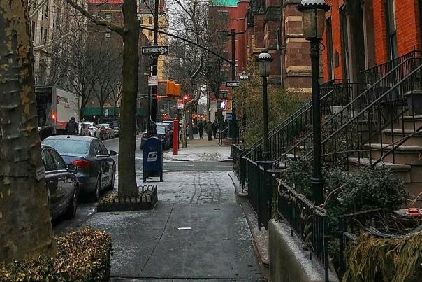 Tree-lined NYC neighborhood street with red brick rowhouses, iron railings, and a stone church spire rising in the background