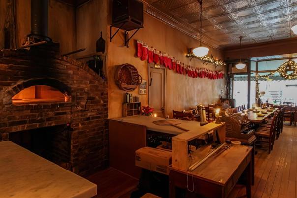 Cozy interior of a New York pizzeria featuring a glowing arched brick wood-fired oven, rustic wooden communal tables, a pressed tin ceiling, and holiday Christmas stockings strung along the wall.
