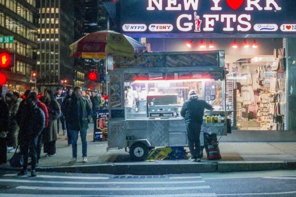 A street food vendor cart glowing under lights at night on a busy Manhattan sidewalk with pedestrians passing by a New York gifts shop