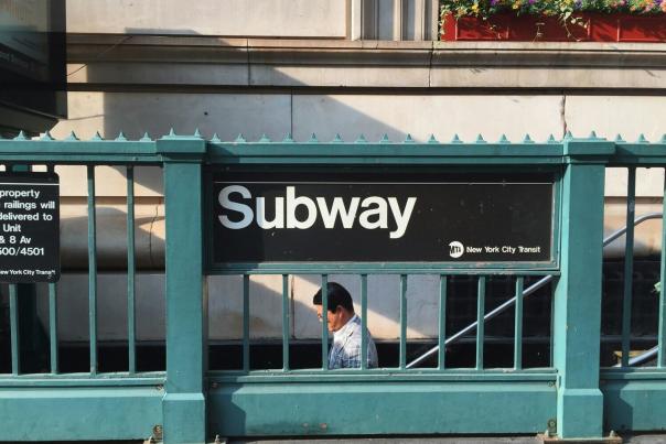 Classic MTA subway entrance with teal iron railings and black Subway sign, a commuter descending the stairs in the background