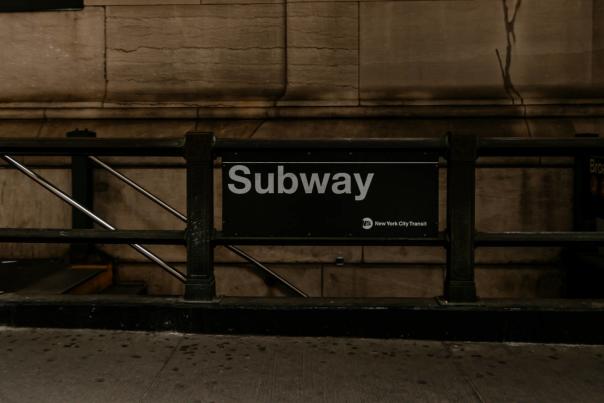 Black “Subway” entrance sign for the New York City subway system mounted on a railing outside a station with stone walls behind it.