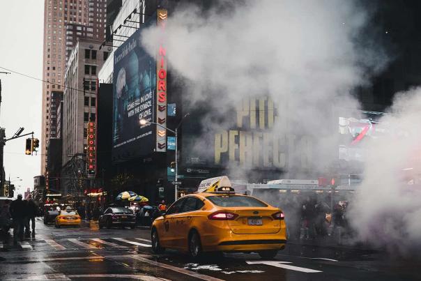 A yellow taxi cab navigates a rainy Times Square intersection as steam rises from the street, with Broadway marquees and neon signs visible in the background