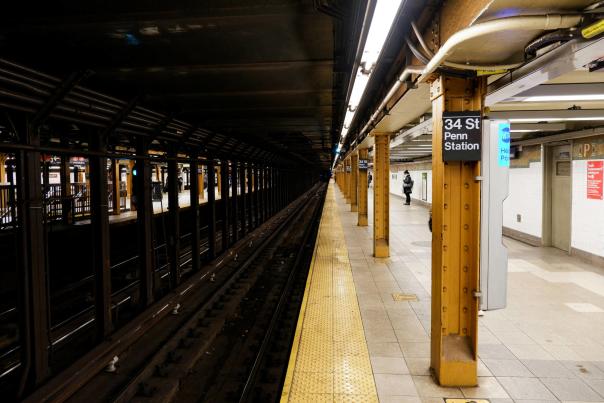 Underground subway platform at 34th Street–Penn Station in New York City with yellow columns, tracks, and commuters waiting along the platform.
