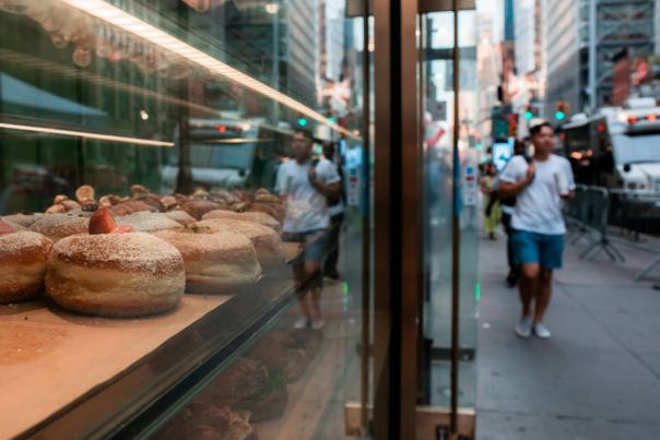 Fresh powdered donuts displayed in a bakery window along a busy New York City sidewalk with pedestrians and traffic in the background.
